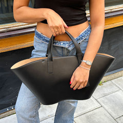 Daniel WellingtonのRiver Bag Blackを手にしたスタイリング例です。デニムとブラックトップスのカジュアルな装いに、ウィングシルエットのブラックレザートートが映え、内側のベージュスエード調素材がちらりと覗くコントラストが魅力的です。