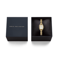 Daniel Wellingtonのギフトボックスに収められたQuadro Mini Arch 3-link White Sunray Gold。ネイビーのパッケージにはブランドロゴが箔押しされた蓋が付き、黒いクッションケースの上にゴールドのスクエア型腕時計が丁寧にセットされています。贈り物にぴったりの上品な仕上がりです。
