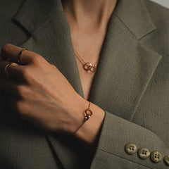 カーキ色のジャケットに合わせたElan Unity Braceletのコーディネートシーン。ローズゴールドのインターロッキングリングが手首でやわらかく輝き、同シリーズのネックレスやリングとのレイヤードスタイルが落ち着いた装いに上品なアクセントを加えています。