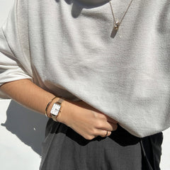 Daniel WellingtonのClassic Bracelet Goldをゴールドのスクエア腕時計やリングと重ね付けしたスタイリング例です。ライトグレーのトップスにダークパンツを合わせたカジュアルなコーディネートに、ゴールドアクセサリーが自然になじんでいます。