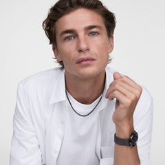 白シャツとTシャツを重ね着した男性モデルが、Tide Mesh Necklace Graphite 1.8を着用しています。グラファイトカラーのメッシュチェーンが襟元からさりげなくのぞき、手首にはダークトーンのDaniel Wellingtonを合わせています。