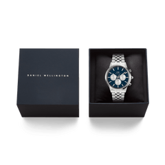 ネイビーのブランドロゴ入りギフトボックスに収められたIconic Chronograph Arch Arctic Panda Silverの俯瞰画像です。ブルーのダイヤルとシルバーブレスレットの腕時計がブラックのクッションに置かれ、贈り物にぴったりの上質なパッケージが確認できます。