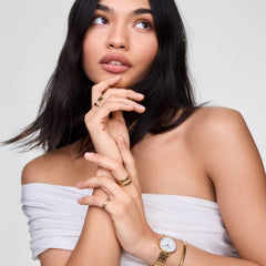 左手首にPetite Roman Numerals Date 5-linkゴールドを着用した女性モデル。ゴールドのリングやブレスレットと重ね付けし、ホワイトトップスに合わせた上品なコーディネートです。