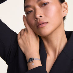 ブラックのジャケットをまとった女性が、Quadro Mini Crystal Zodiac Sterlingを手首に着用したスタイリングです。クリスタルベゼルに縁取られたブラックマザーオブパールの文字盤と、星座チャームが揺れるシルバーメッシュストラップが、上品な装いに自然になじんでいます。