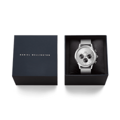 Daniel Wellingtonのネイビーカラーのブランドボックスに収められたIconic Chronograph Mesh Silverのギフトパッケージです。箱の蓋にはブランドロゴが箔押しされ、ブラックのクッショニングの上にシルバーメッシュベルトのクロノグラフ時計が丁寧にセットされています。