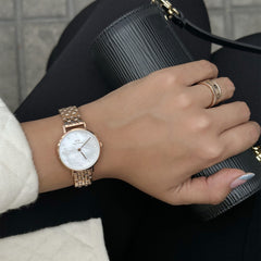 Petite Lumine Melroseを着用した手元のスタイリング写真です。白蝶貝のダイヤルとローズゴールドの5リンクブレスレットが、黒のバッグとコーディネートされ、ローズゴールドのリングとの重ねづけで上品な印象を演出しています。