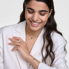 白いジャケットを着た女性が、Daniel WellingtonのAudrey Ring Mother of Pearl Goldを薬指にはめ、同コレクションのグリーンストーンのブレスレットやネックレス、イヤリングとゴールドトーンでコーディネートしたスタイリング例です。