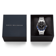 ダニエルウェリントン Iconic GMT Link Black Blue Ceramic Bezel Silverが、ネイビーのブランドロゴ入りギフトボックスに収められています。ブラックのクッションの上にシルバーリンクブレスレットの腕時計が丁寧にセットされており、贈り物に最適な上質なパッケージです。