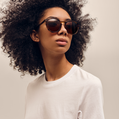 ダニエルウェリントンのArch Brown Demiを着用した女性のモデル写真です。べっ甲柄のブラウンデミのラウンドフレームとアンバーレンズが、白いトップスとのコーディネートに自然に馴染んでいます。