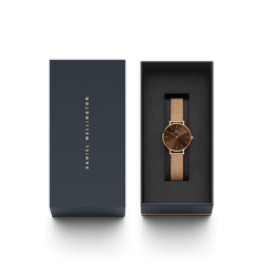 Daniel Wellington Petite Amber Rose Goldをネイビーの専用ギフトボックスに収めた状態の俯瞰画像です。箱の蓋にはゴールドのブランドロゴが刻印され、内側のブラッククッションにはアンバーブラウンの文字盤とローズゴールドのメッシュストラップをあしらった腕時計が丁寧にセットされています。