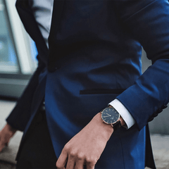 ネイビーのスーツに白シャツを合わせた男性の手元から、Daniel Wellington Classic Sheffield Black Rose Goldがのぞくビジネスシーンの着用イメージです。ブラックの文字盤とローズゴールドのケースが、スーツスタイルに上品に馴染みます。