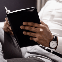 白シャツにグレーのパンツを合わせた男性が、ソファでくつろぎながら読書を楽しむひととき。手首にはDaniel Wellington Classic Sheffieldのブラックレザーストラップが、自然な装いに溶け込んでいます。