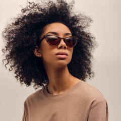 Daniel Wellington Lynx Bio-Acetateのブラウンデミ キャットアイサングラスを着用した女性モデル。べっ甲柄のフレームとアンバーレンズが、ベージュトーンのカジュアルなコーディネートに自然になじみます。