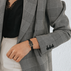 グレンチェックのジャケットにブラックのインナーを合わせたオフィススタイルに、Daniel Wellington Quadro Pressed Ashfieldを着用した女性の手元です。ブラックのメッシュストラップとローズゴールドケースが、モノトーンコーディネートに自然に馴染んでいます。