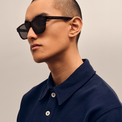 ネイビーのポロシャツを着用した男性が、Daniel Wellington Ambler Bio-Acetate Dark Havanaのサングラスをかけた横顔のクローズアップです。テンプル部分にはゴールドのブランドロゴが刻印されています。