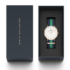 Classic Warwick ローズゴールドが、ネイビーのブランドギフトボックスに収められています。箱の蓋にはゴールドの Daniel Wellington ロゴが箔押しされ、ネイビーとグリーンの NATO ストラップを装着した腕時計が、黒いクッションケースの上に丁寧にセットされています。