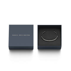 ダニエルウェリントン Classic Bracelet Graphiteのパッケージ画像。ネイビーのブランドロゴが入ったギフトボックスの蓋が開かれ、黒いクッショントレーの上にグラファイトカラーの細身なカフブレスレットが収められています。